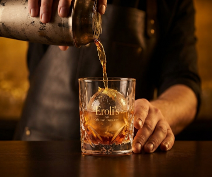 Barkeeper gießt einen Cocktail mit Eiskugel und Eroli's Logo in der Bar am Platz an der Schmiede ein